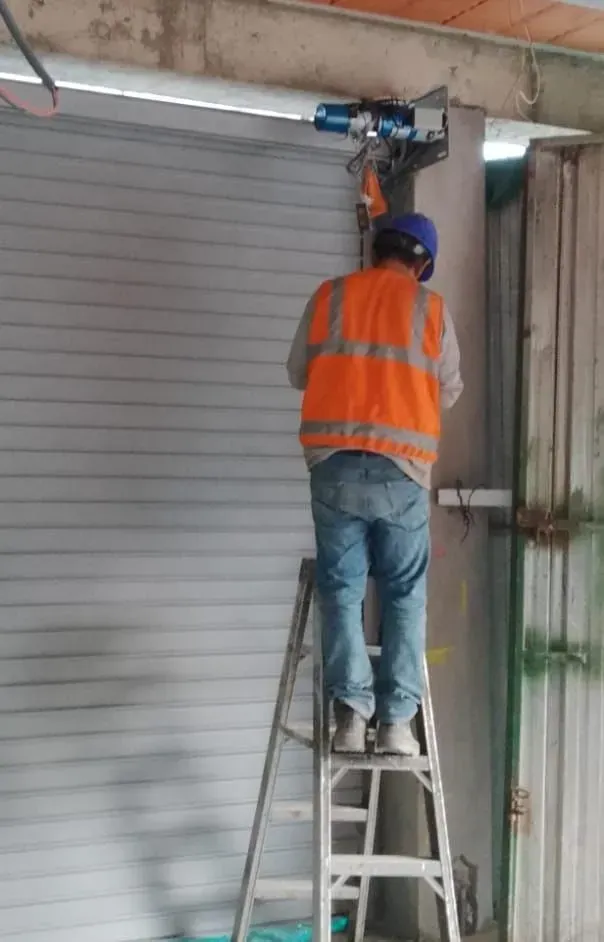 Un trabajador subido a una escalera instala un abrepuertas de garaje motorizado azul sobre una puerta metálica enrollable.