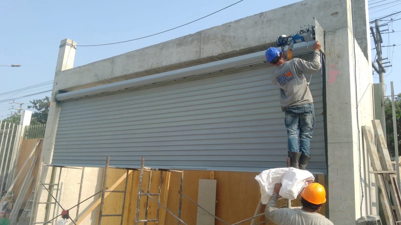 Un trabajador subido a una escalera instala una persiana enrollable de metal gris en el interior de un edificio industrial.
