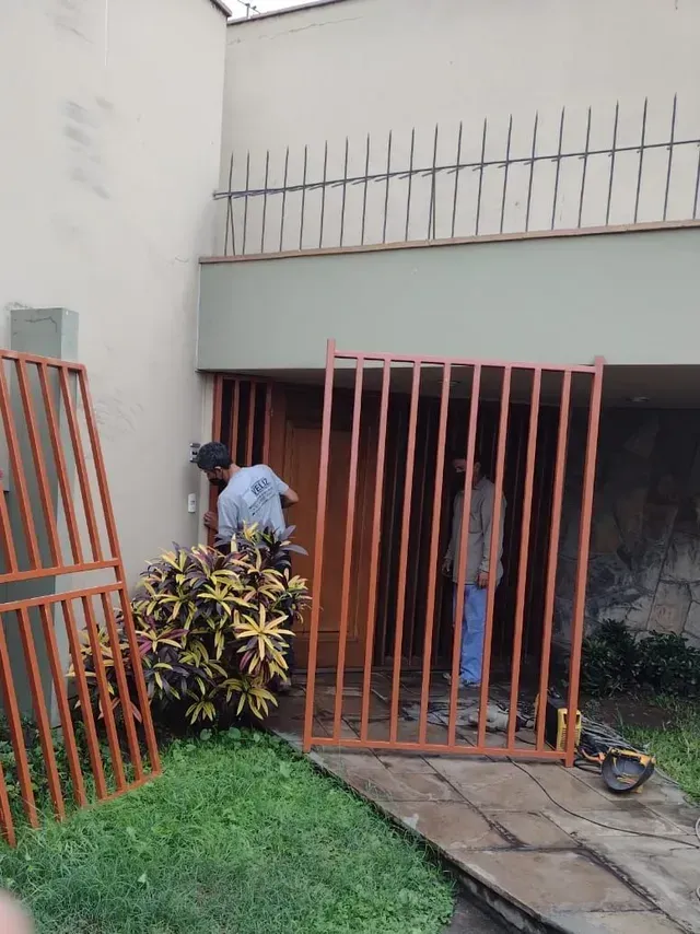 Dos personas detrás de unas rejas metálicas de color naranja, en el exterior de un edificio, con un arbusto frondoso y césped delante.