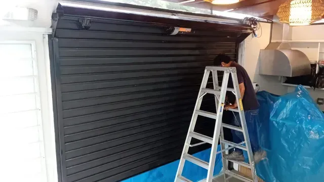 Una persona sube a una escalera para ajustar una puerta de garaje enrollable negra en un espacio de trabajo desordenado.