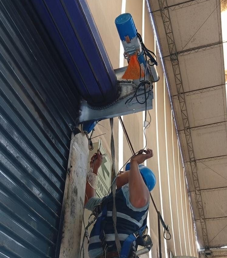Dos personas detrás de unas rejas metálicas de color naranja, en el exterior de un edificio, con un arbusto frondoso y césped delante.
