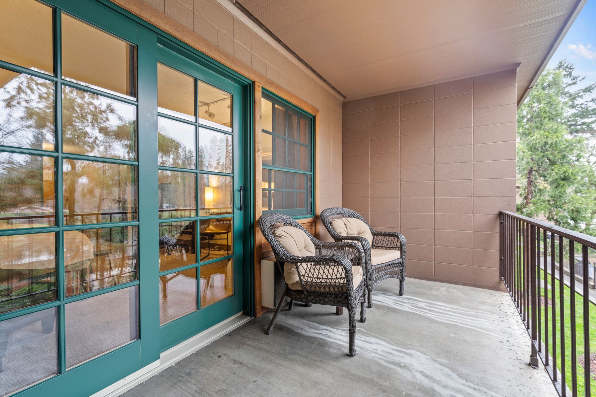 Covered balcony with teal French doors, two wicker chairs, and a railing overlooking trees.