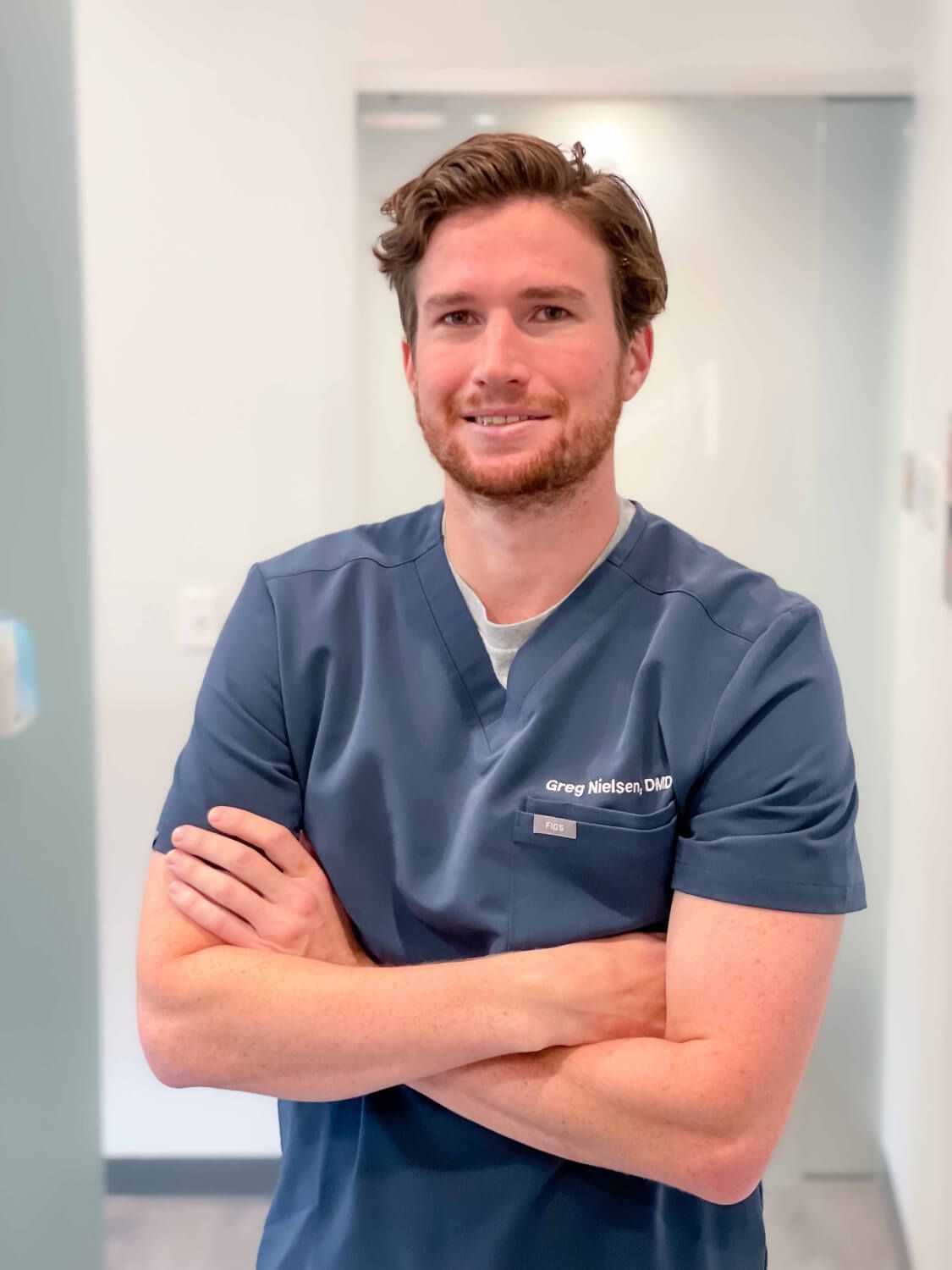 Man in blue scrubs smiles, arms crossed, in a brightly lit hallway.