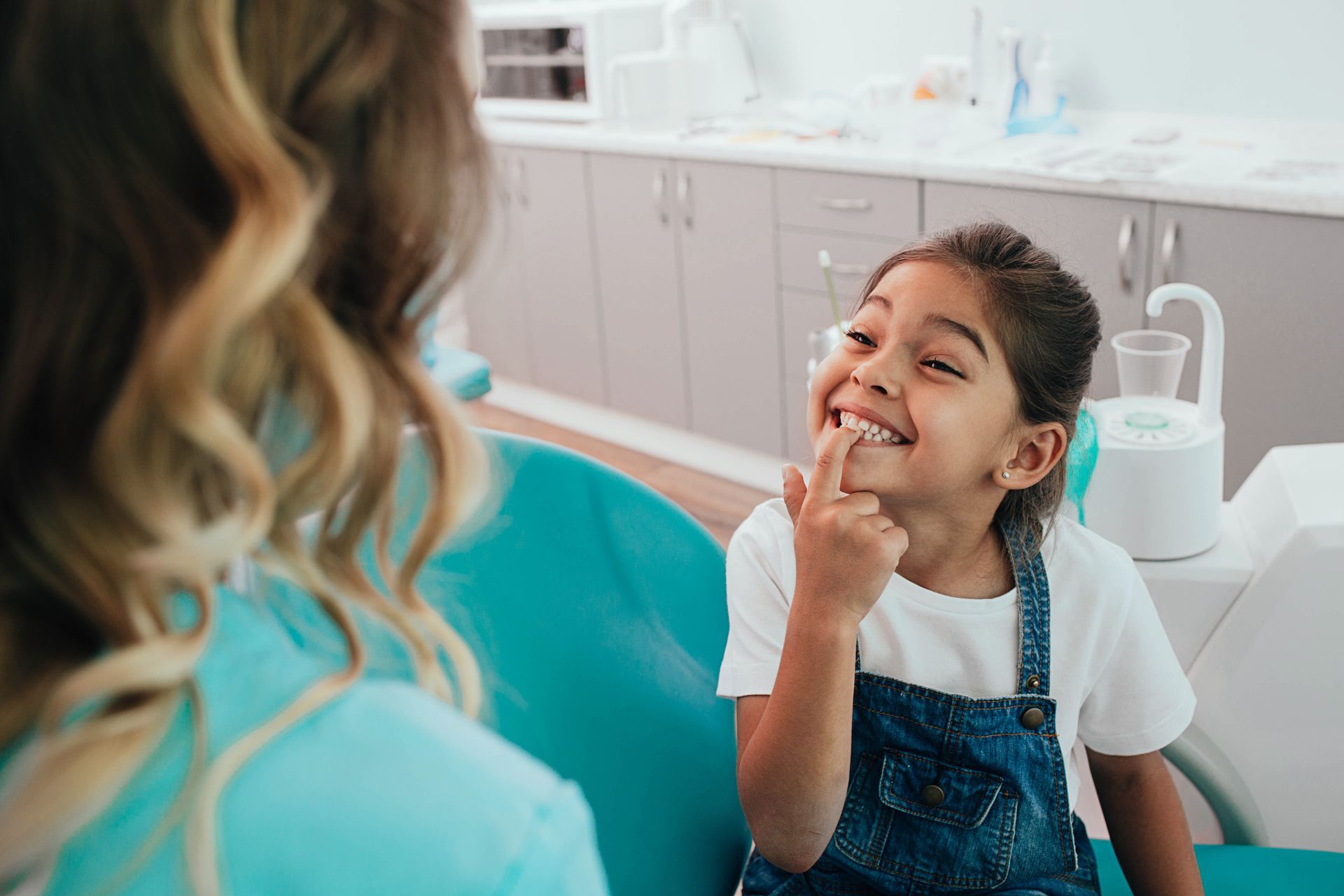 Young girl in dental chair pointing at her teeth, smiling. Dentist in background.