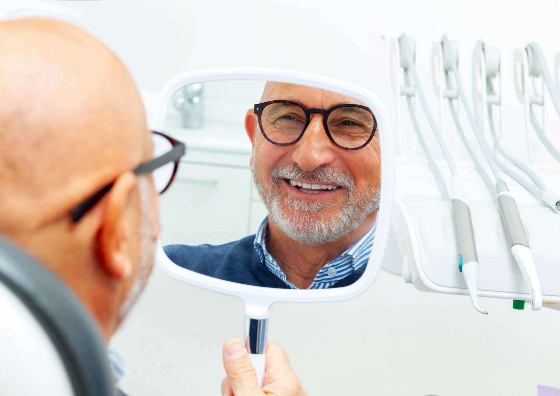Bald man with glasses smiles while looking at his teeth in a dental mirror.