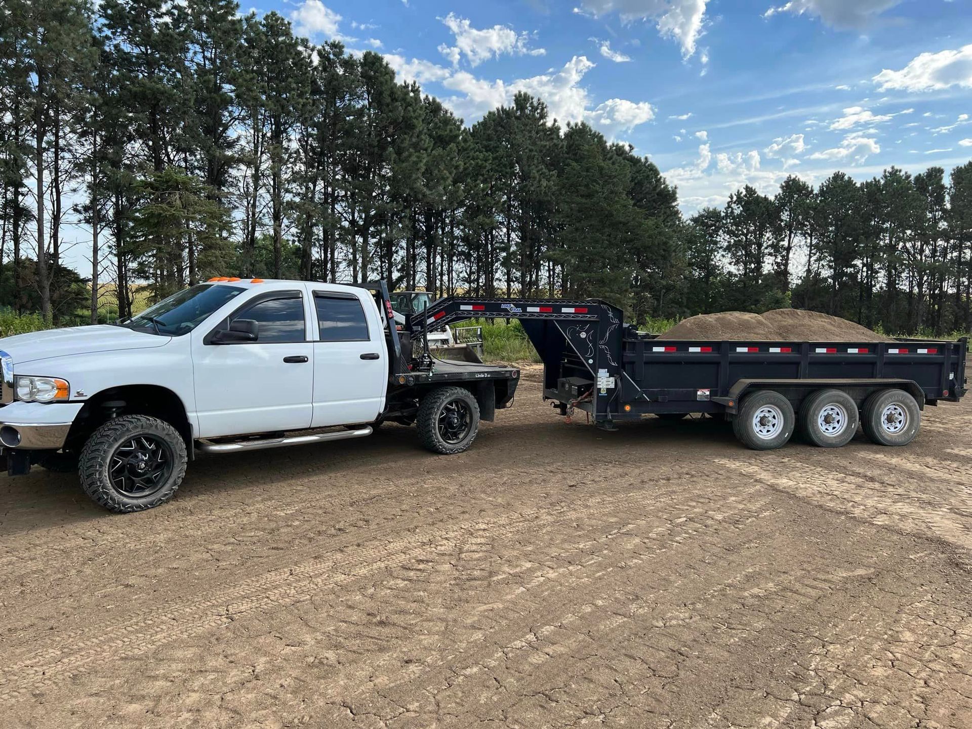 A white truck is towing a dump trailer in a dirt field.