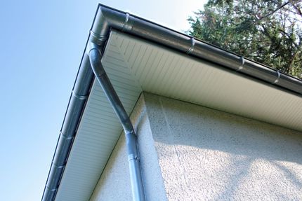 Coin d'un bâtiment avec gouttières grises, descente de gouttière et soffite blanche, sur fond de ciel bleu.