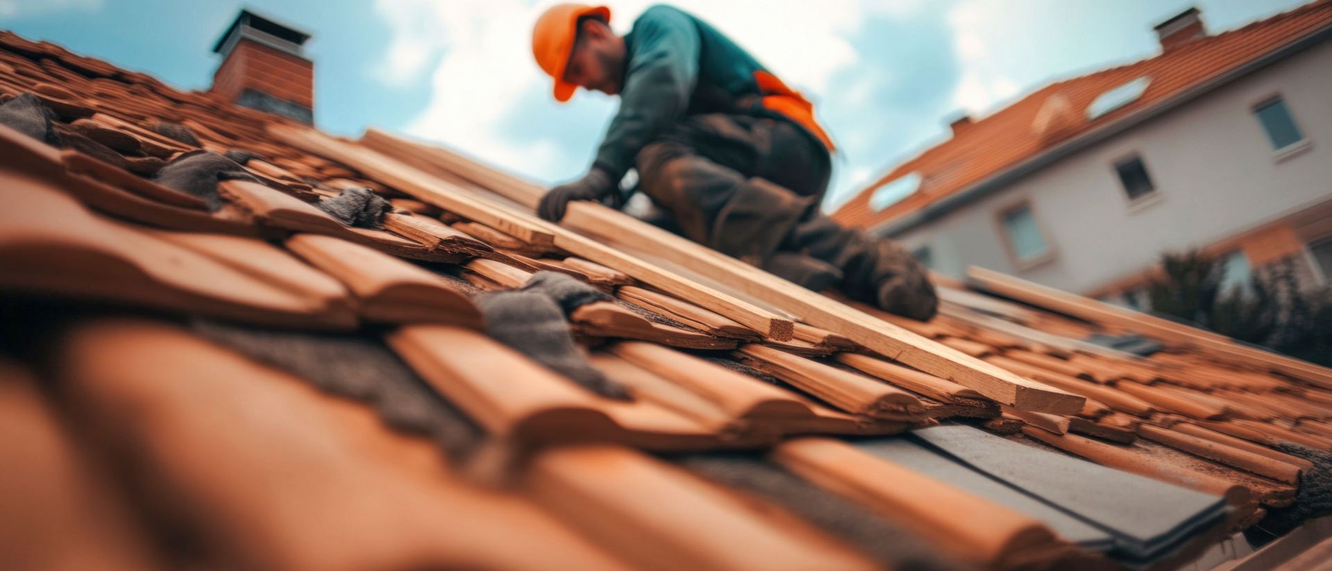 Un couvreur en tenue de sécurité effectue des réparations sur un toit de tuiles ; d'autres bâtiments sont visibles en arrière-plan.