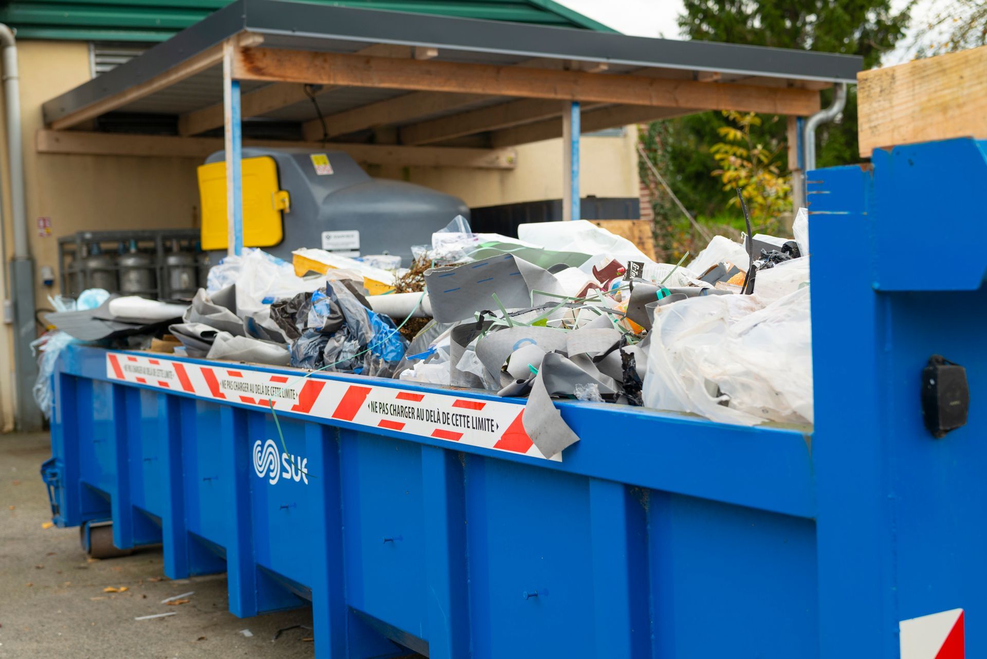 Conteneur à ordures bleu rempli de déchets, sous un toit en bois.