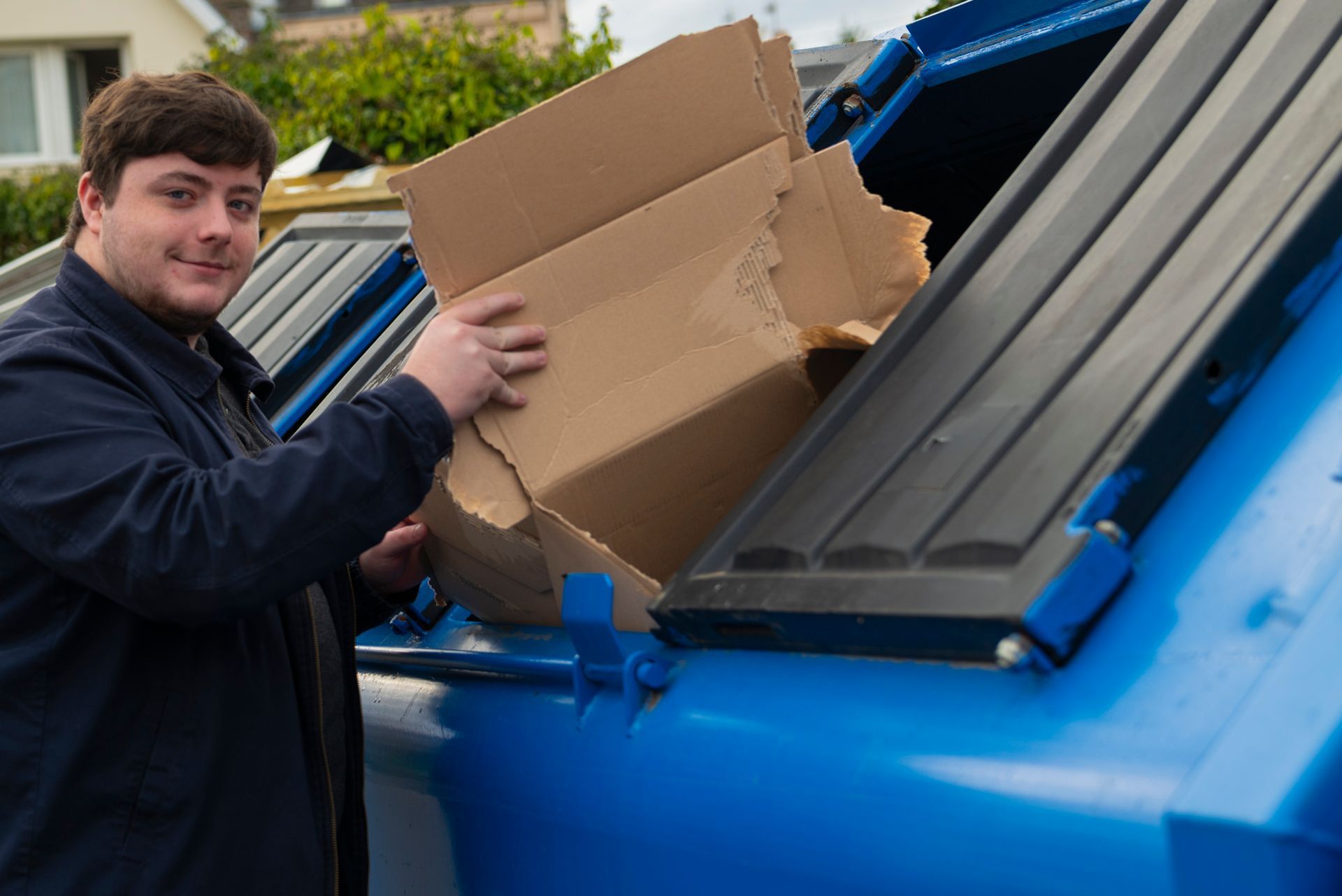 Un homme dépose un carton dans un bac de recyclage bleu. Il sourit, à l'extérieur.