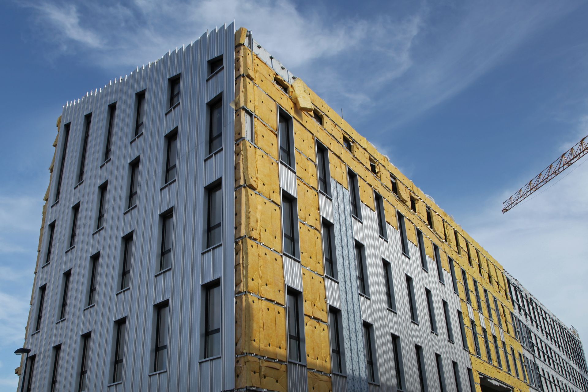 Bâtiment en construction, avec un revêtement métallique gris, une isolation jaune et un ciel bleu.