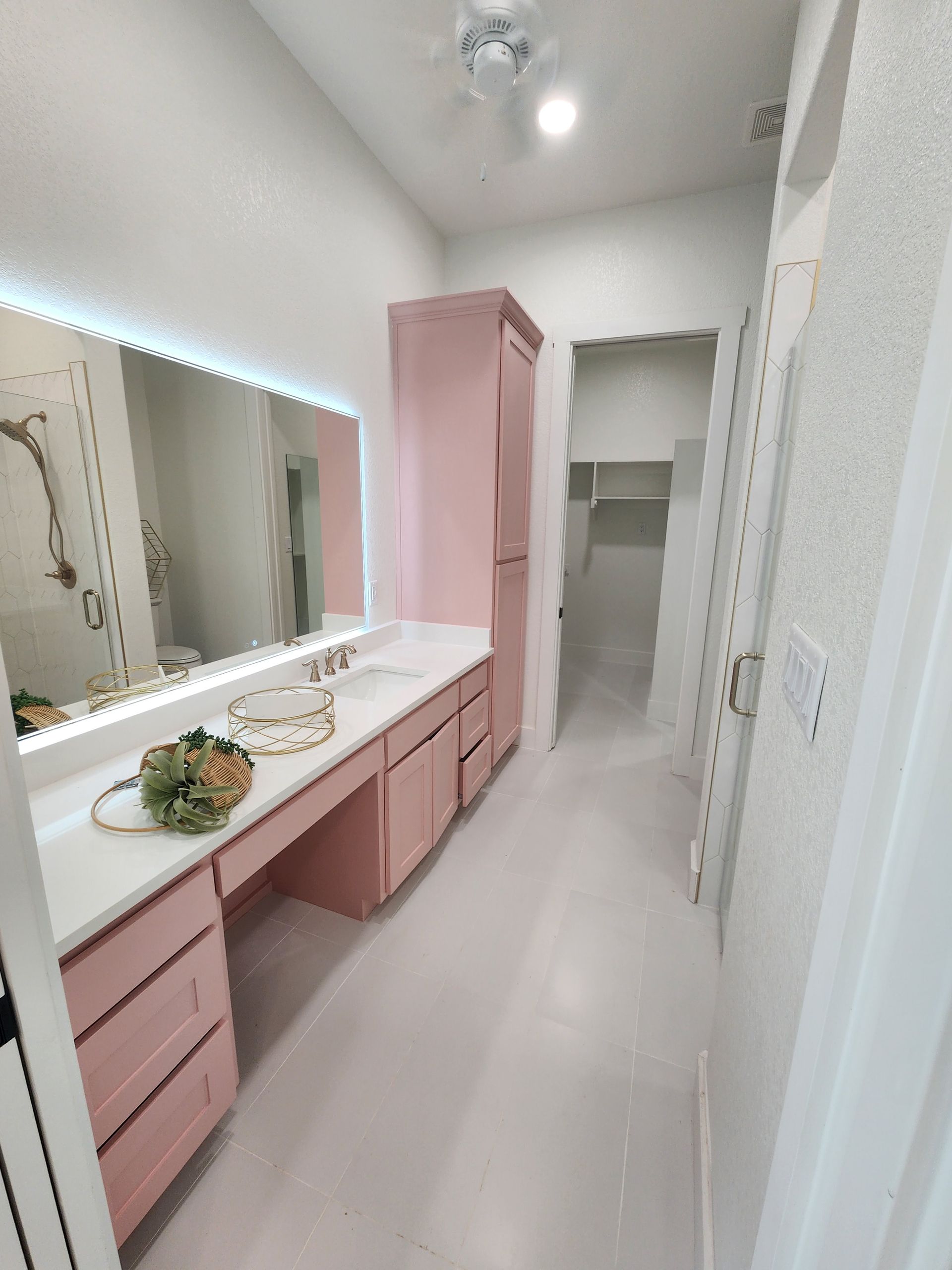 Narrow pink-and-white bathroom vanity with mirror, sink, and open shelving in a bright hallway-like room