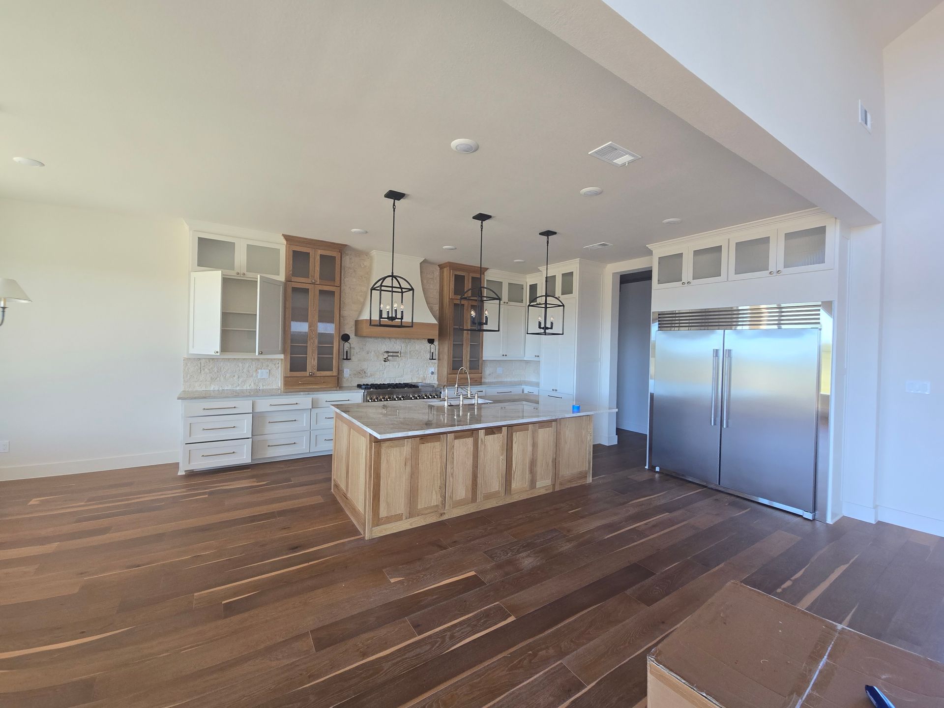 Empty modern kitchen with wood floors, white cabinets, large island, and stainless steel refrigerator