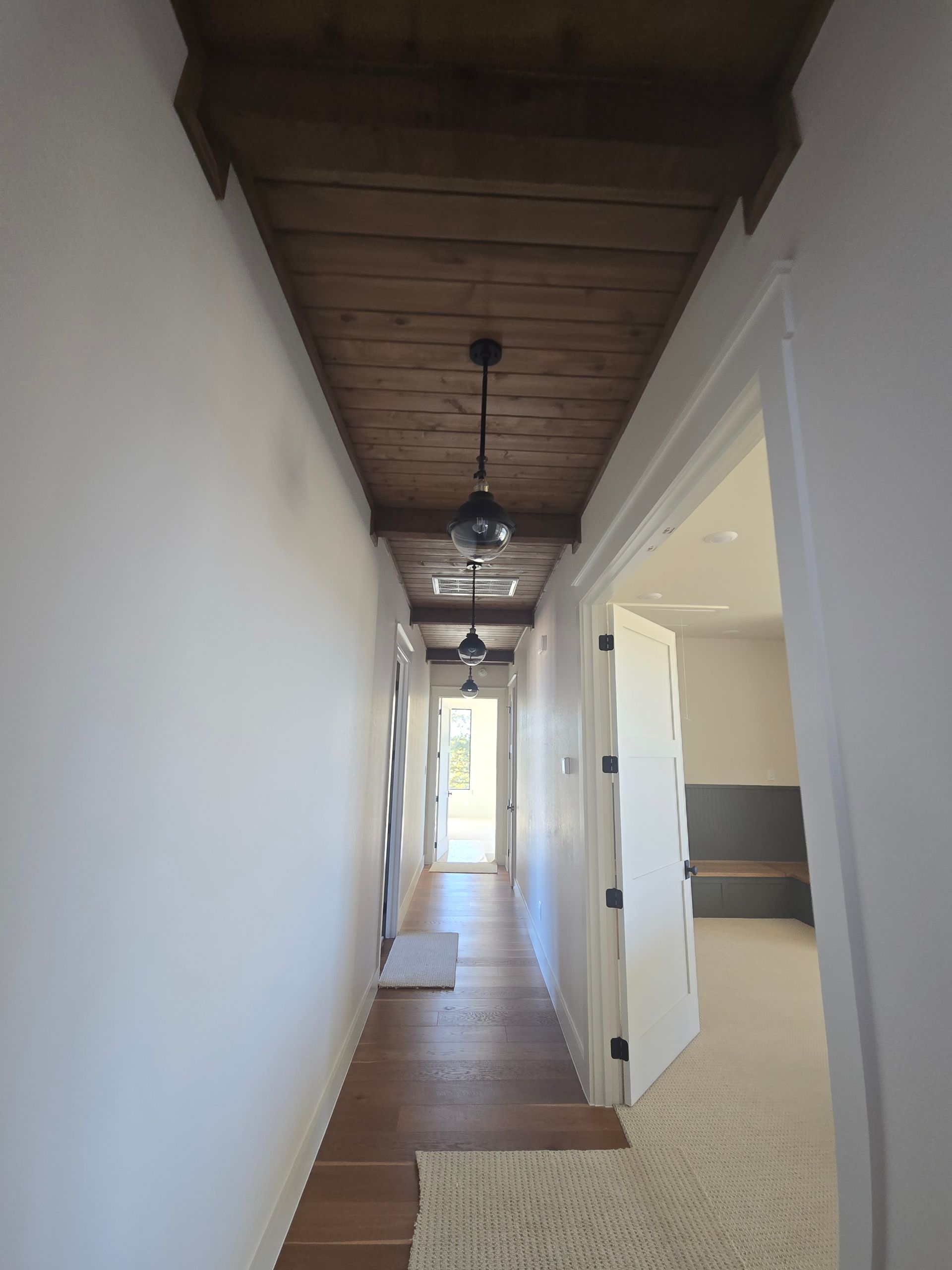 Narrow hallway with wood ceiling, hanging lights, and open doors leading to a bright room.