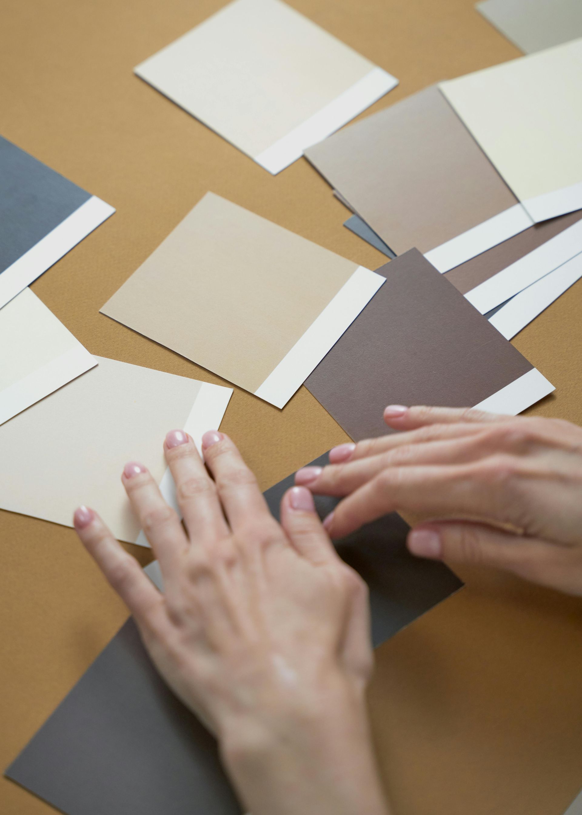 Hands arranging several rectangular color swatches in shades of brown, beige, and blue on a tan surface.