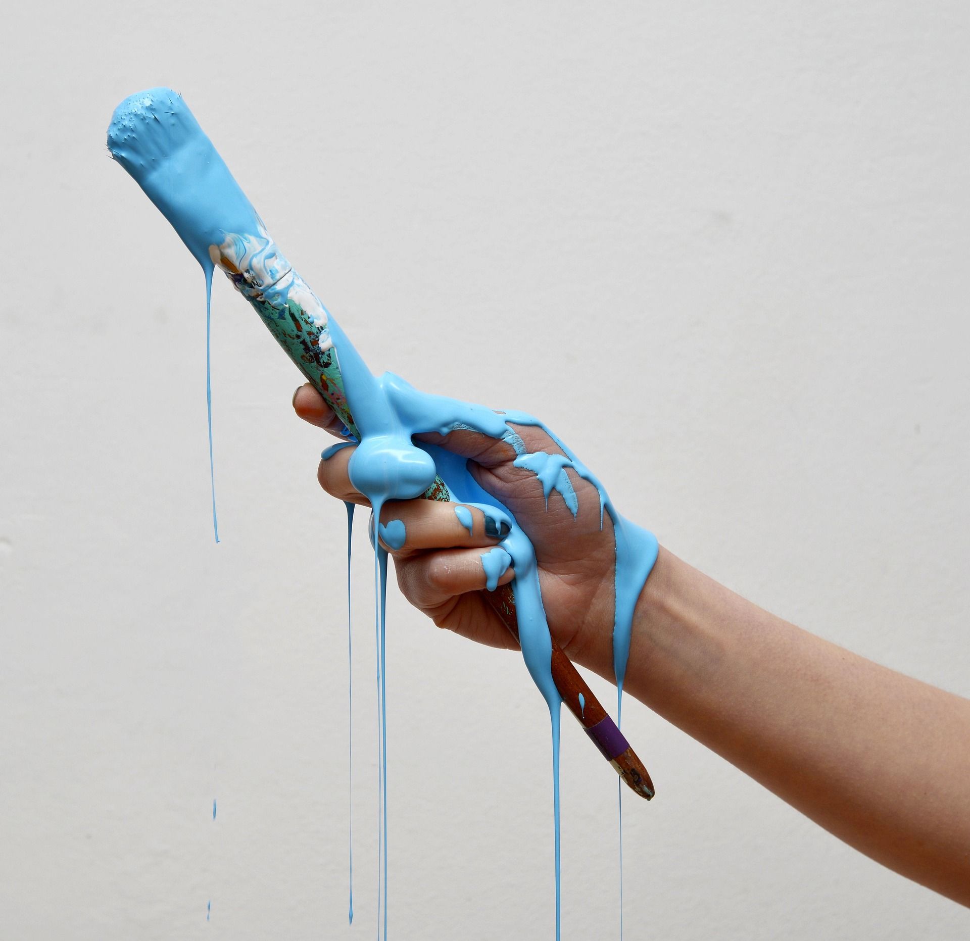 A hand holds a paintbrush while thick, bright blue paint drips down the brush, fingers, and wrist against a white wall.