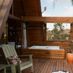 A wooden deck features a white jacuzzi, a green wooden chair with a floral pillow, and a small potted orange plant.