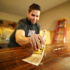 A person using a brush to apply a dark stain to a wooden surface in a workshop with a ladder in the background.