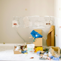 A room under renovation with a beige wall, electrical outlets, a cardboard box, paint supplies, and flooring covered.