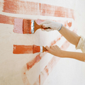 Two hands in light-colored work gloves paint horizontal stripes of orange paint onto a light-colored wall with brushes.