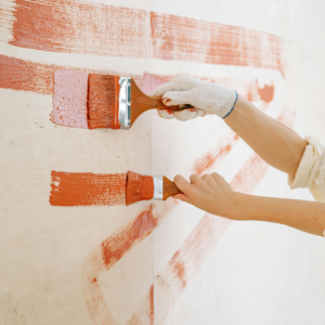 Two hands in light-colored work gloves paint horizontal stripes of orange paint onto a light-colored wall with brushes.