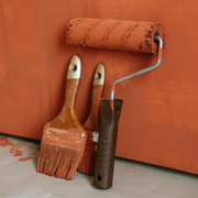 A paint roller and two paintbrushes covered in terracotta-colored paint rest against a wall painted the same shade.