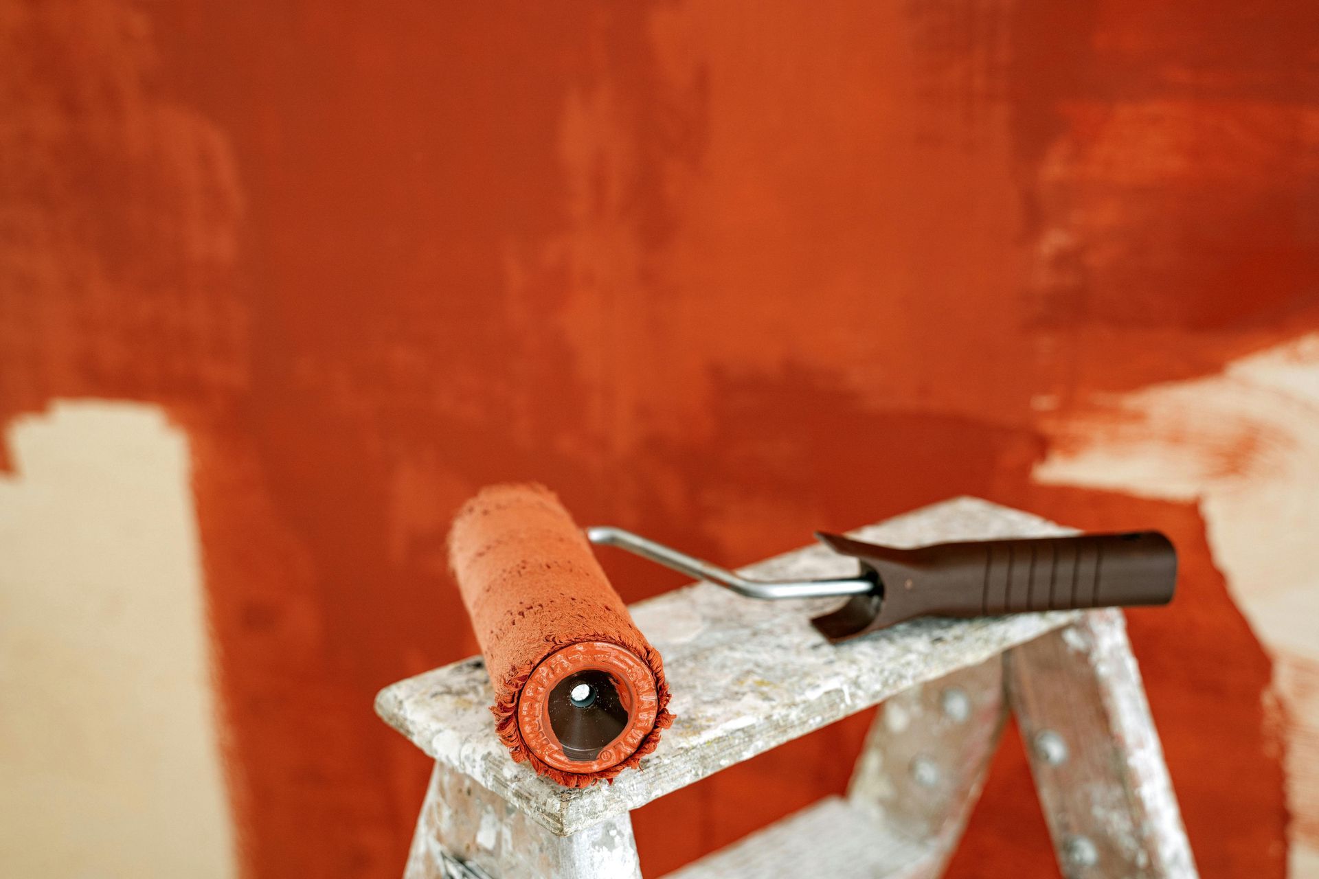 A paint roller with orange paint rests on the metal step of a ladder against a partially painted orange wall.