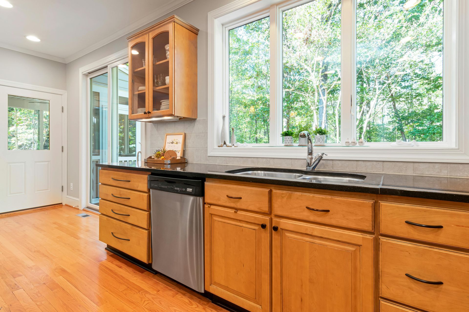 A bright kitchen with honey-colored wood cabinets, a stainless steel dishwasher, a window over the sink, and wood floors.