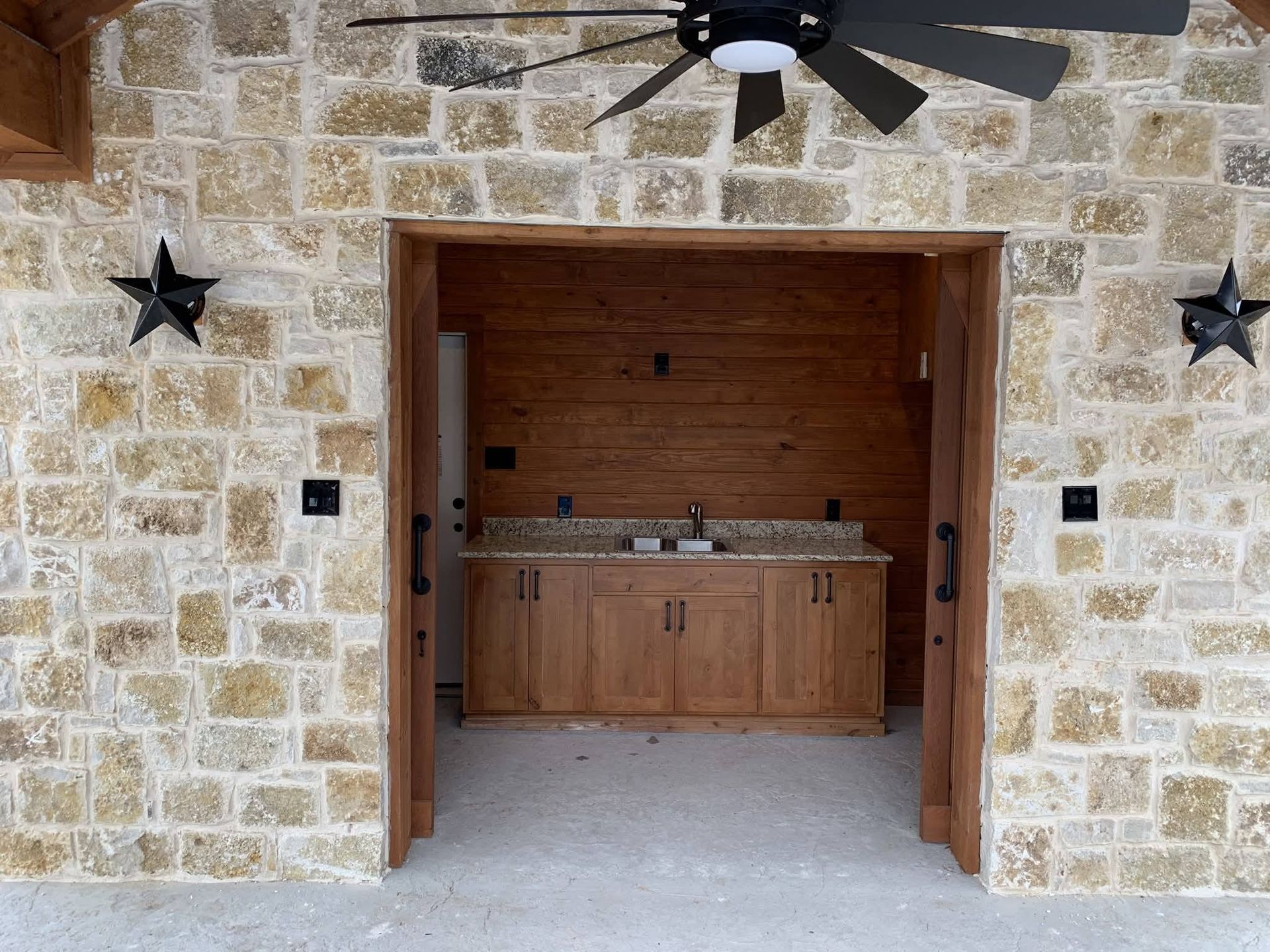 Stone patio with open wooden double doors leading to an outdoor kitchen sink and counter beneath a ceiling fan