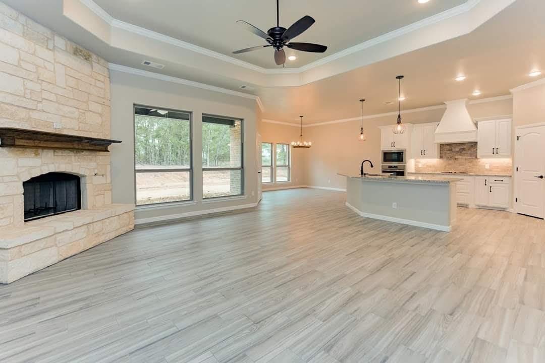 Spacious modern living room with stone fireplace, large windows, white kitchen, and light wood floors