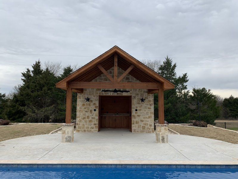 Stone pavilion with wood beams and a central doorway beside a blue pool under a cloudy sky