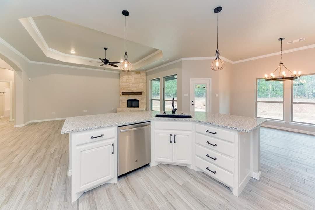 Bright open kitchen with white cabinets, granite island, stainless dishwasher, and large windows overlooking a living area