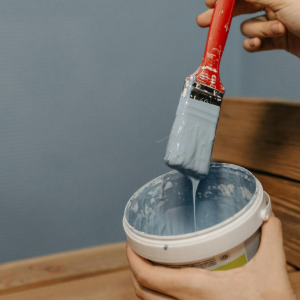 A hand holds a paintbrush dripping light blue paint over an open paint container.