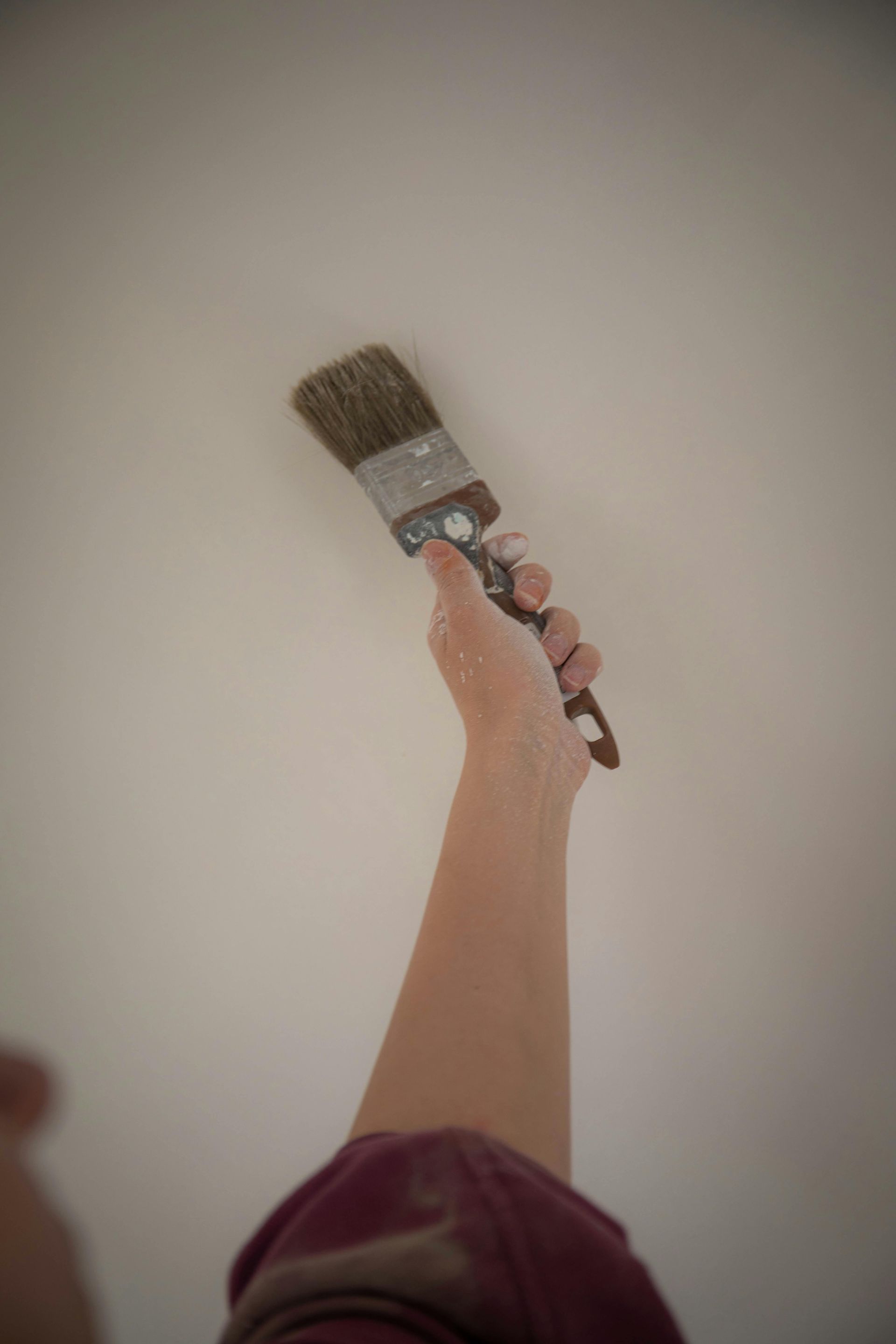 A person's arm reaches up to paint a plain white ceiling with a brush.