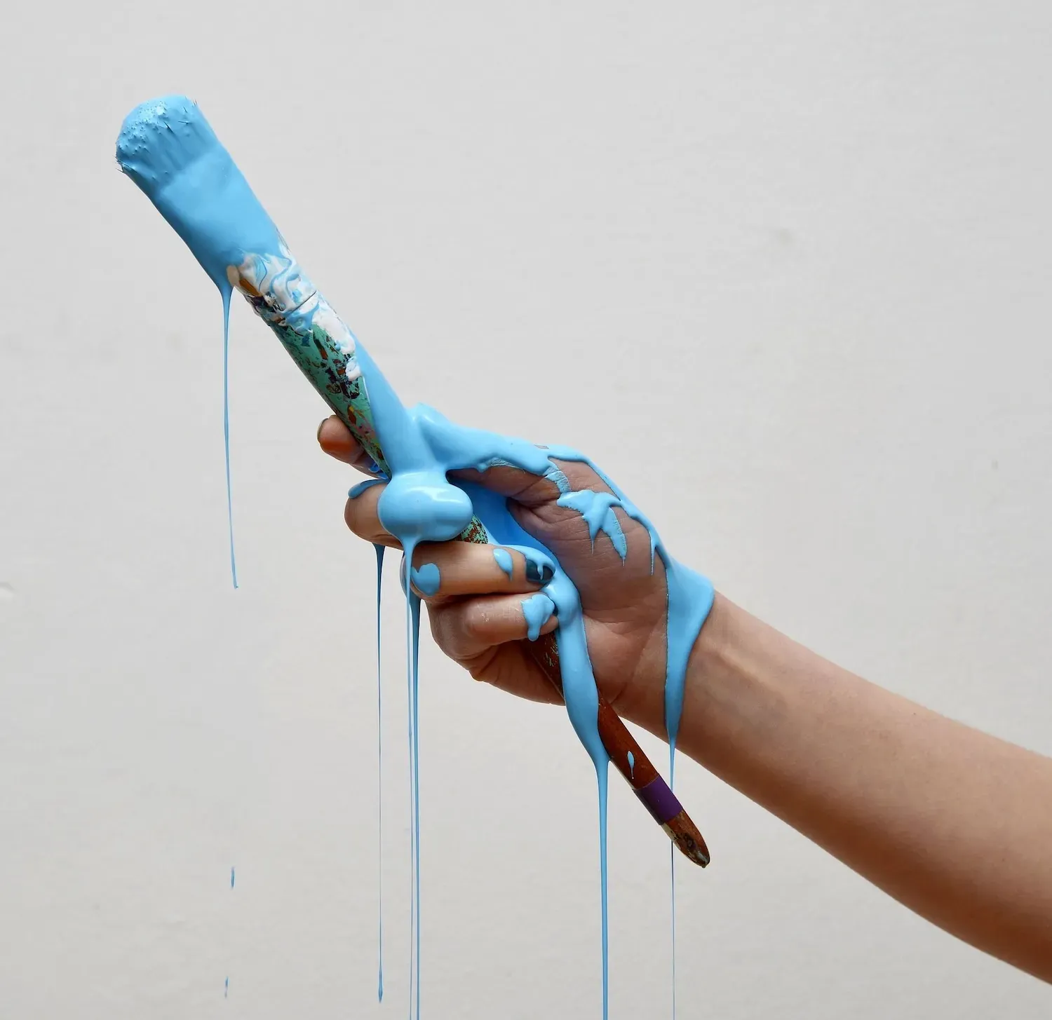 A hand holds a paintbrush while thick, bright blue paint drips down the brush, fingers, and wrist against a white wall.