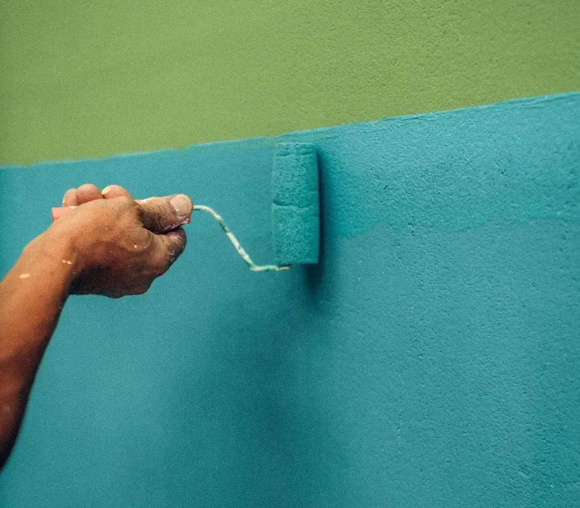A hand uses a paint roller to apply teal paint to a textured wall below a band of green paint.