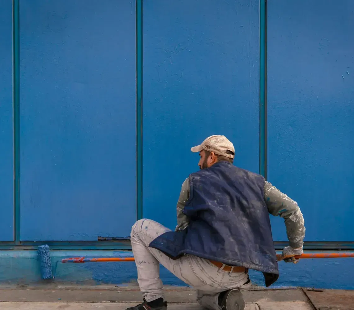 A person in a blue vest and light pants squats while using a long-handled roller to paint a bright blue wall.