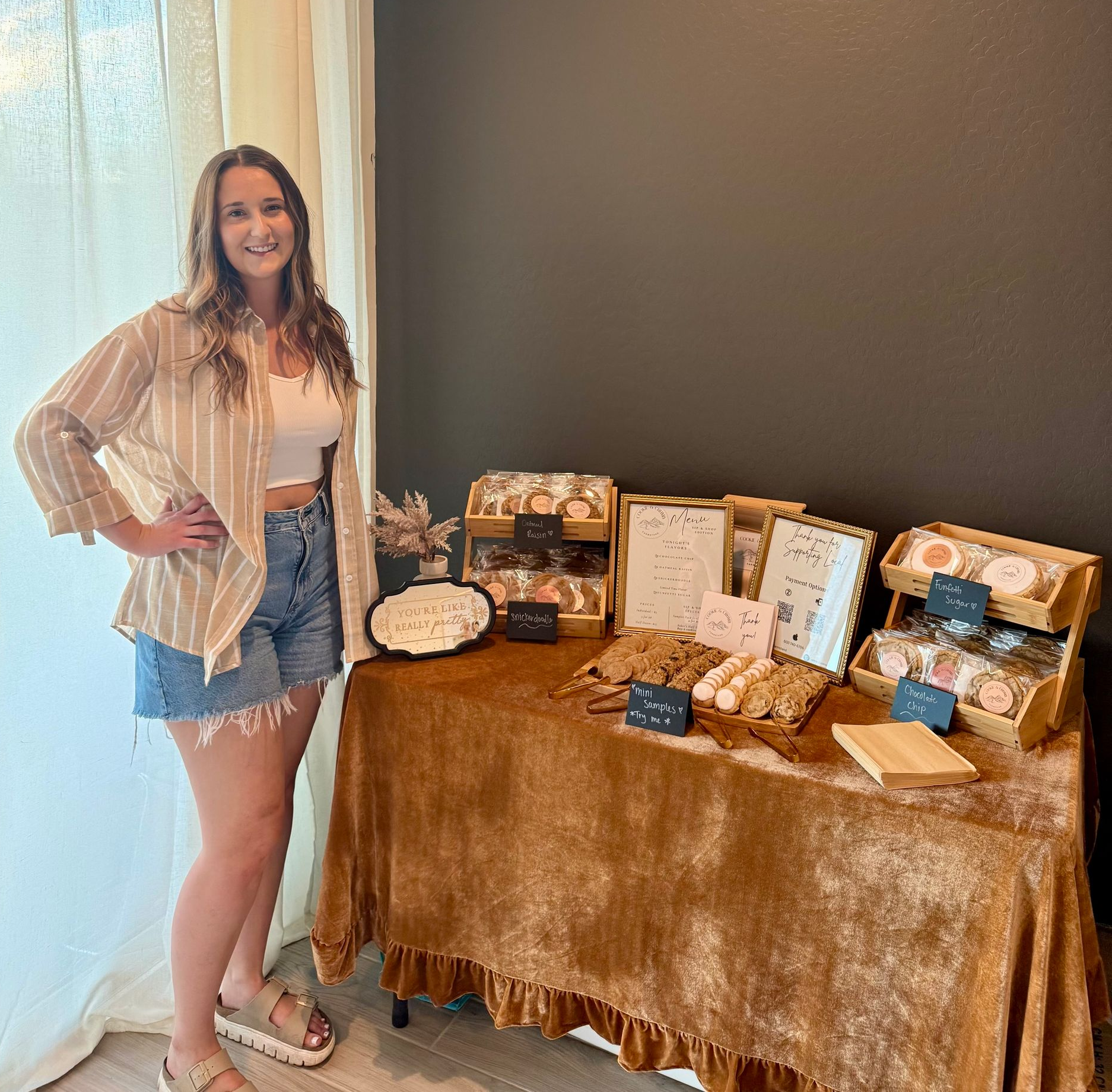 Molly stands at a table displaying baked goods. She wears shorts, a white top, and a striped shirt, brown backdrop.