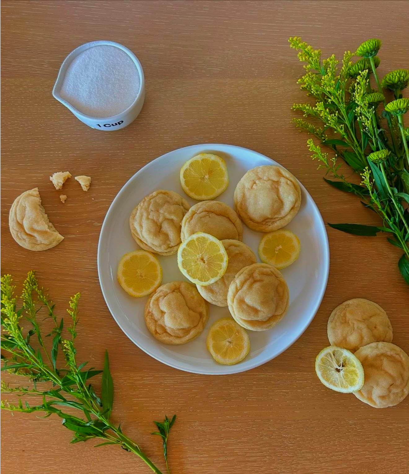 Lemon cookies and slices on a white plate with sugar, flowers, and wood background.