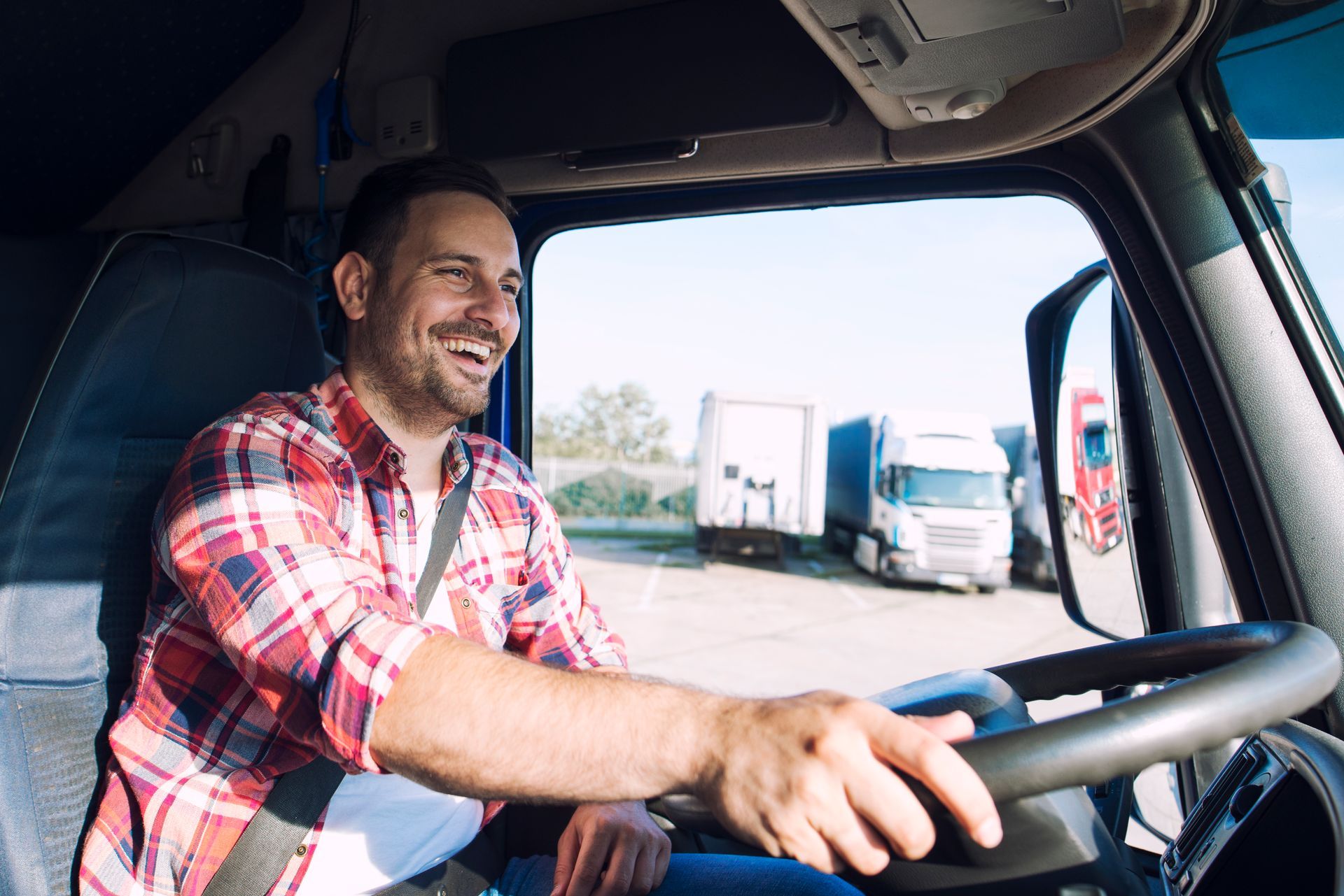Smiling truck driver in red plaid shirt driving a semi with trucks visible outside the window