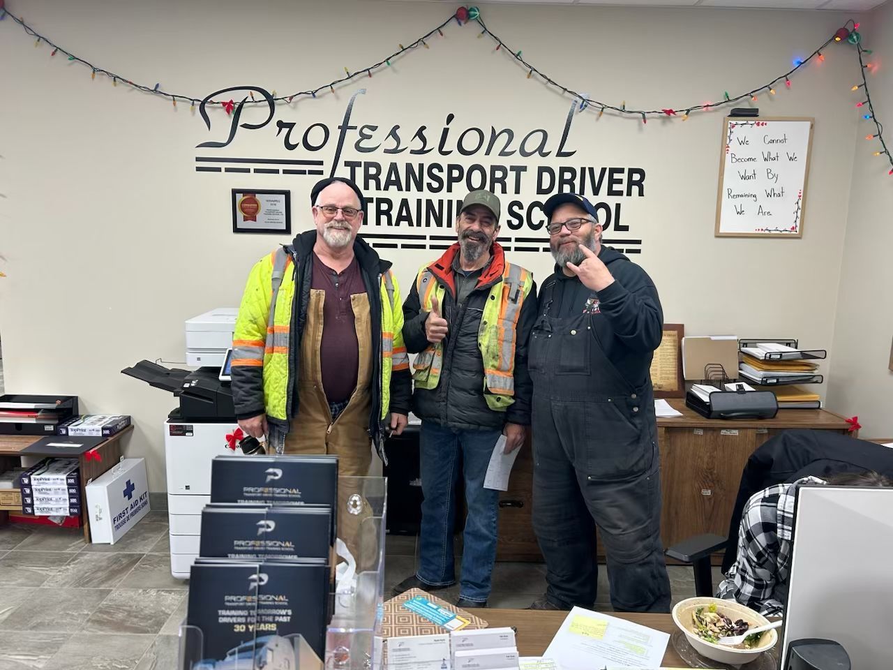 Three people stand in an office under a sign for the Professional Transport Driver Training School.