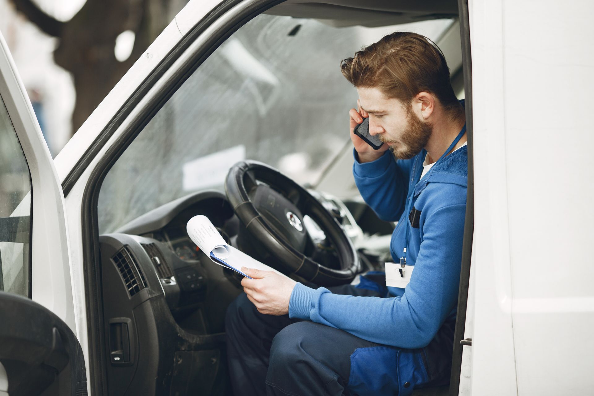 Man in blue shirt sitting in a white car, talking on a phone while reading a document