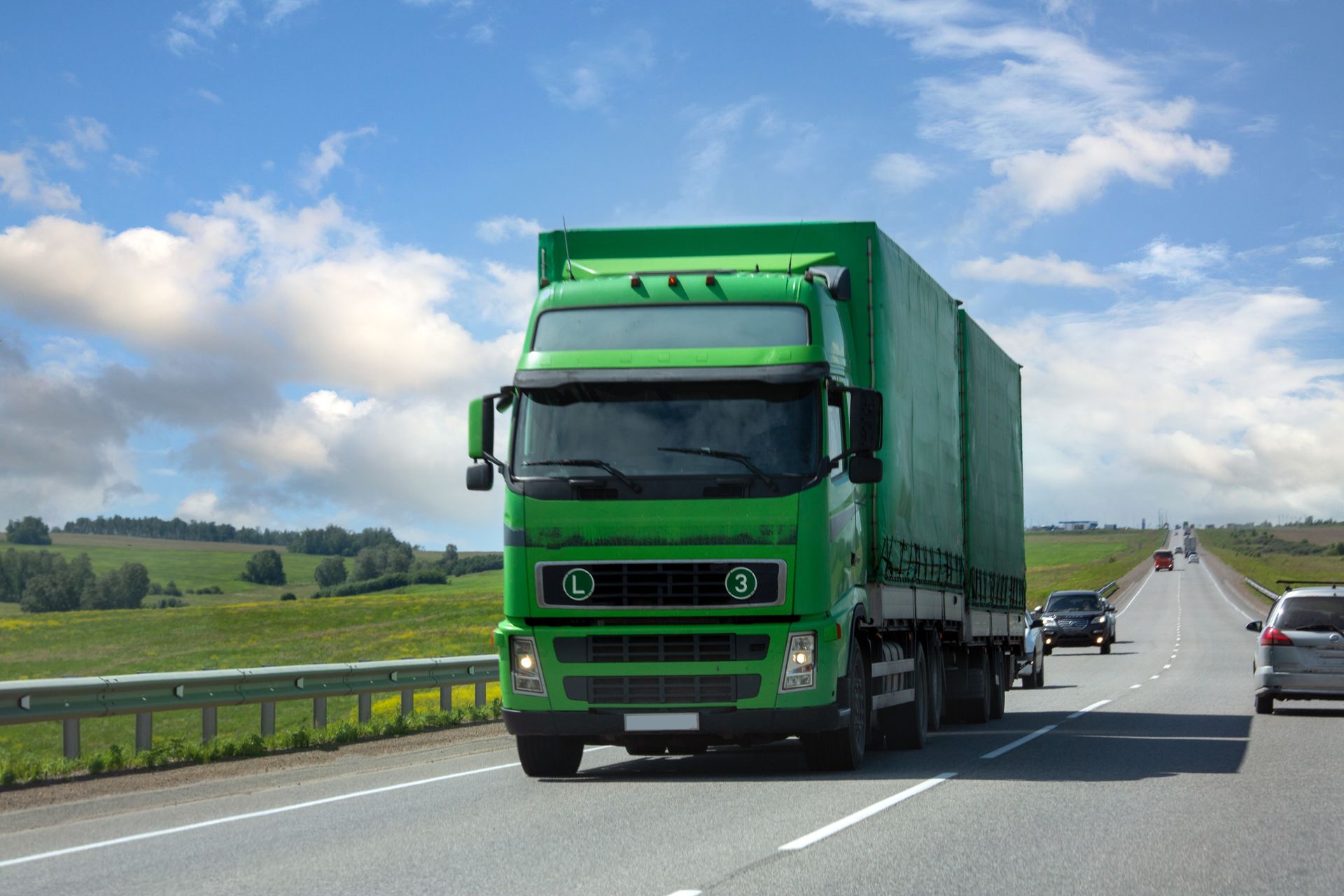 Green semi-truck driving on a highway with open fields and a cloudy sky