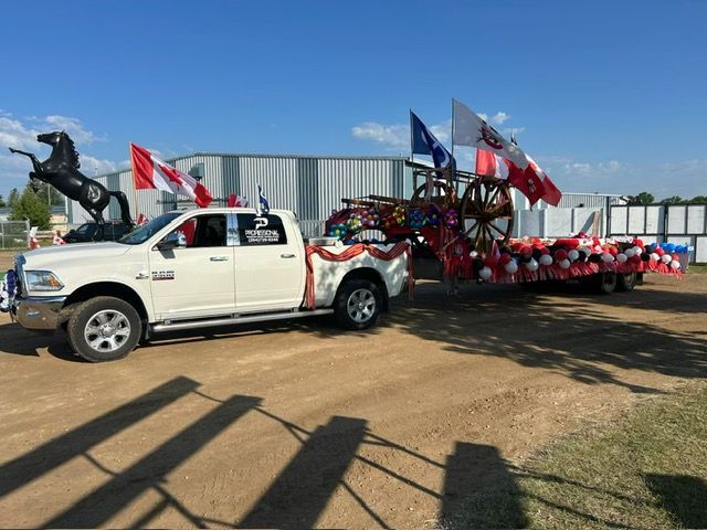 White pickup towing a red-and-black parade float with flags on a dirt road, statue visible in the background