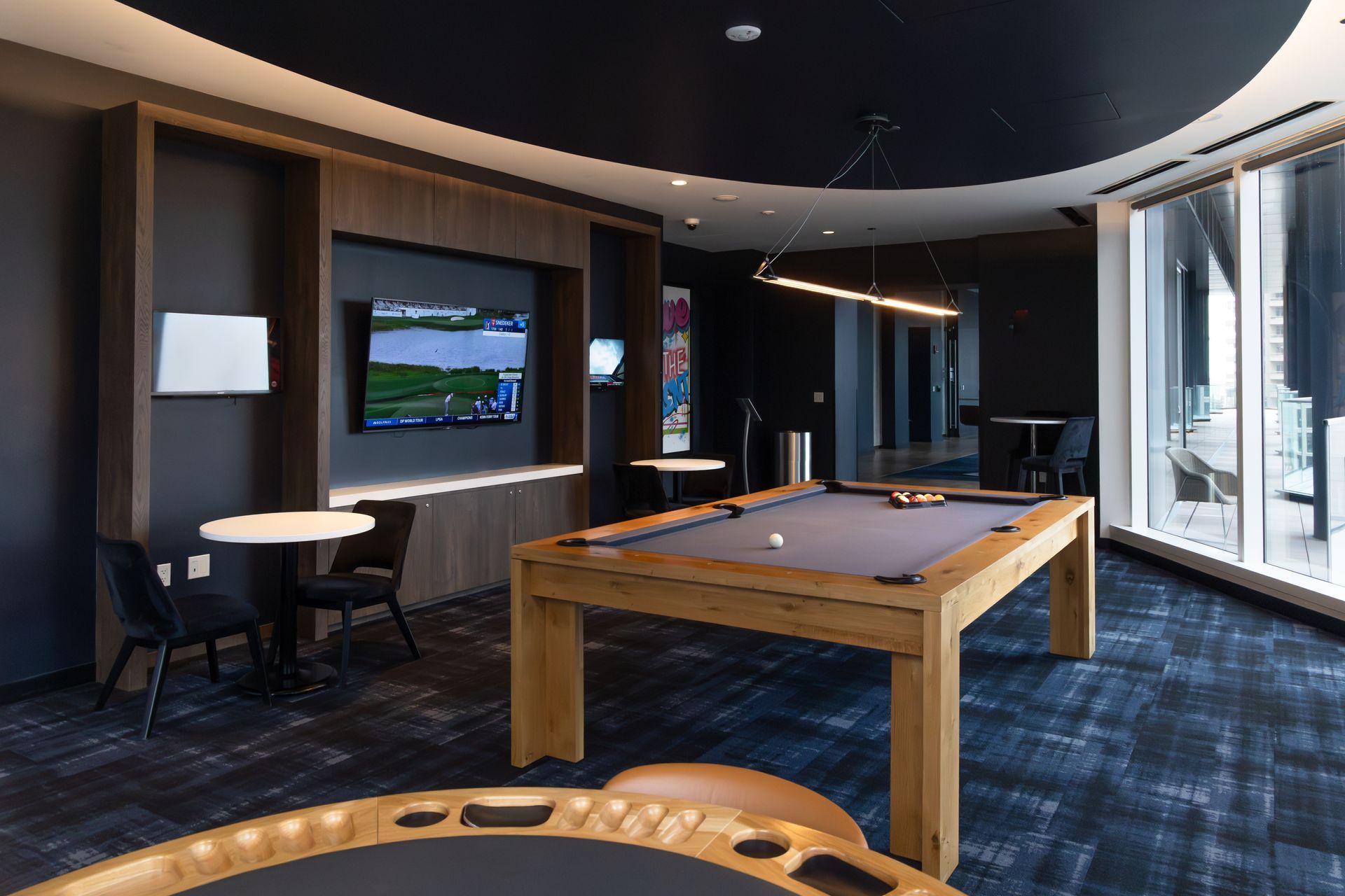 Game room with pool table, TV, and small tables. Dark walls, patterned carpet.