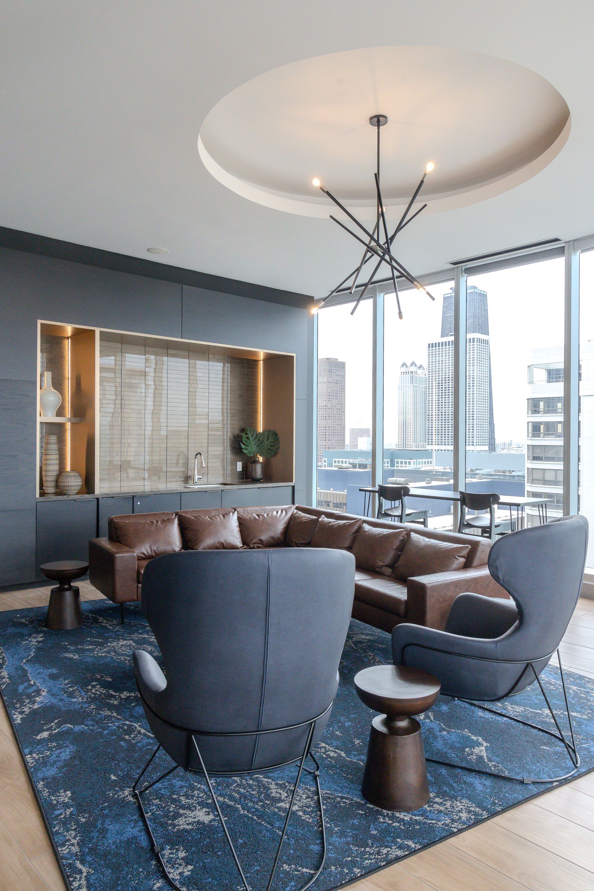 Modern lounge with blue rug, brown sofa, gray chairs, and floor-to-ceiling city view.