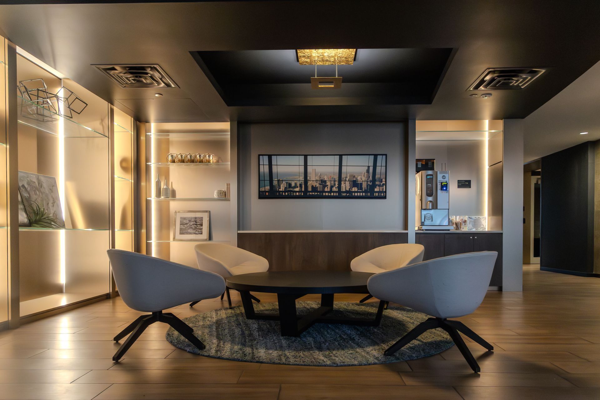 Lounge area with round table, four chairs, and illuminated cabinets. Gray walls, artwork, coffee machine.