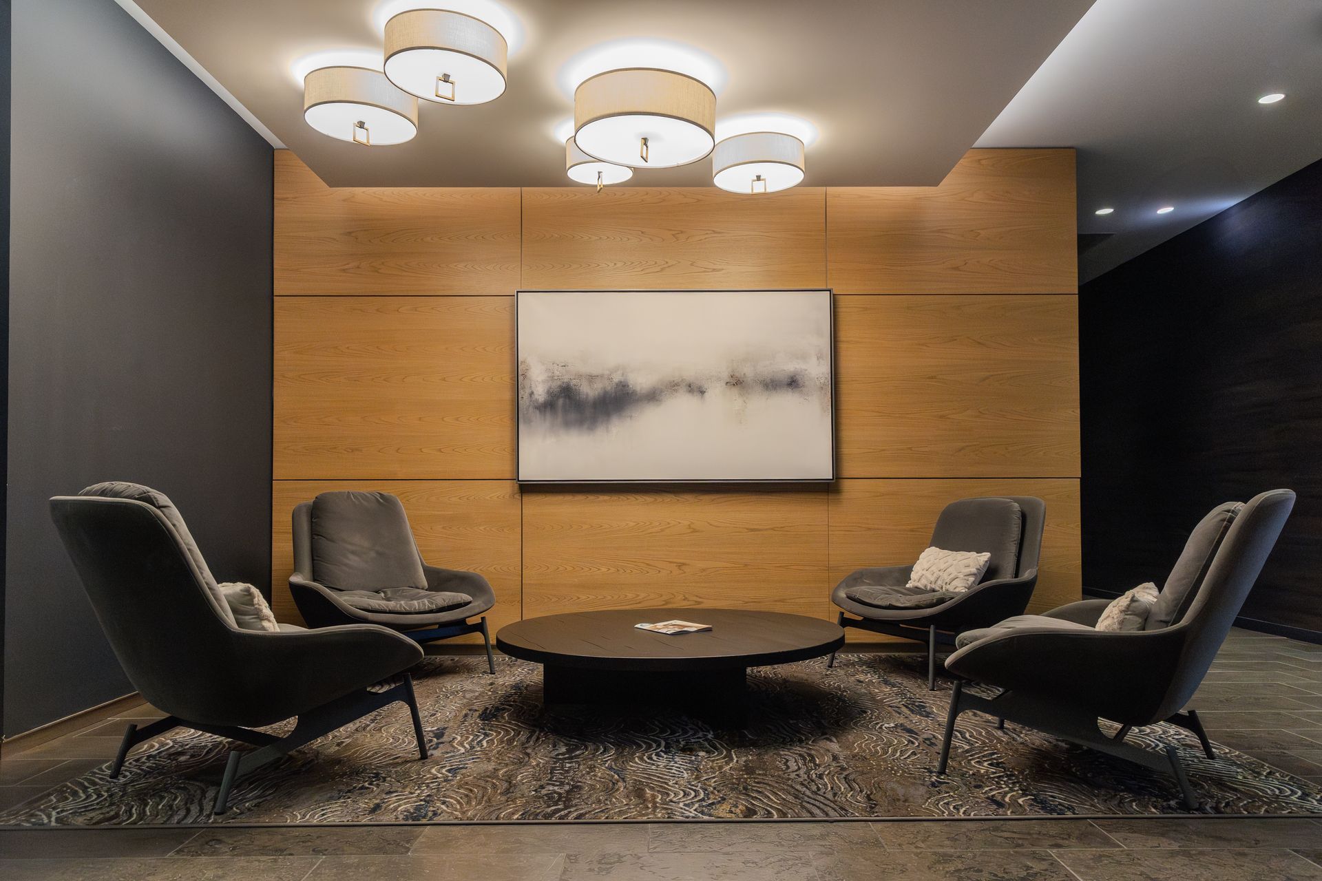 Lounge area with four gray armchairs around a round coffee table. Abstract art on wood paneling, lit by ceiling lights.