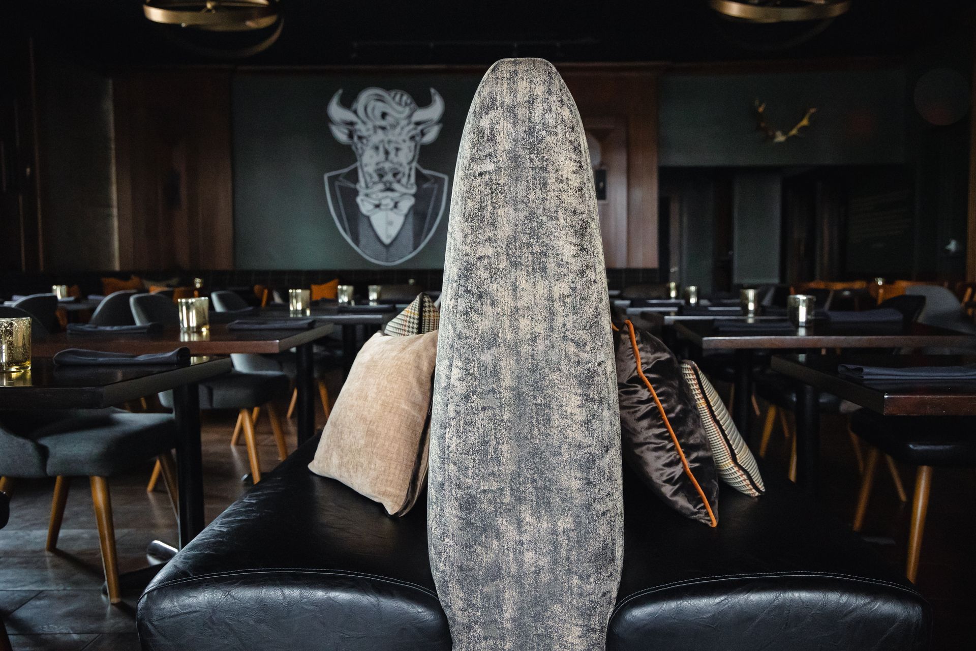 Dark restaurant interior with a large wooden sculpture, leather couch, tables, and a bison logo on the wall.