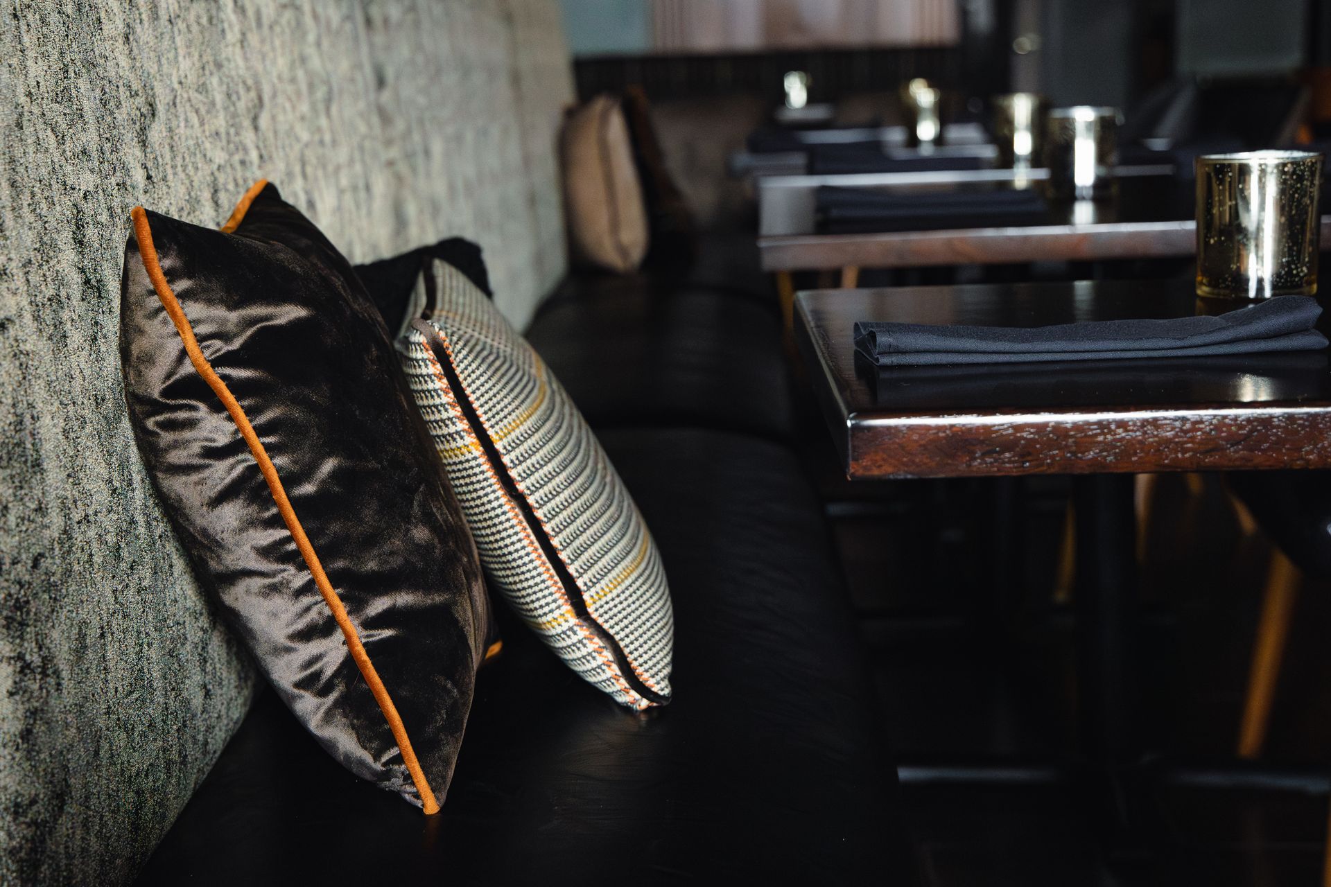 Black leather bench with pillows against a textured wall, next to tables set with place settings in a restaurant.